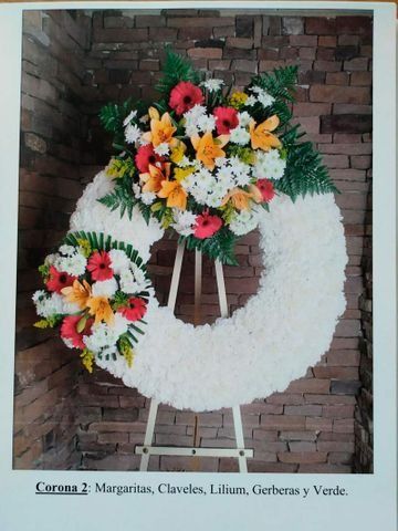 Corona funeraria blanca con flores de colores sobre un soporte contra una pared de ladrillos.