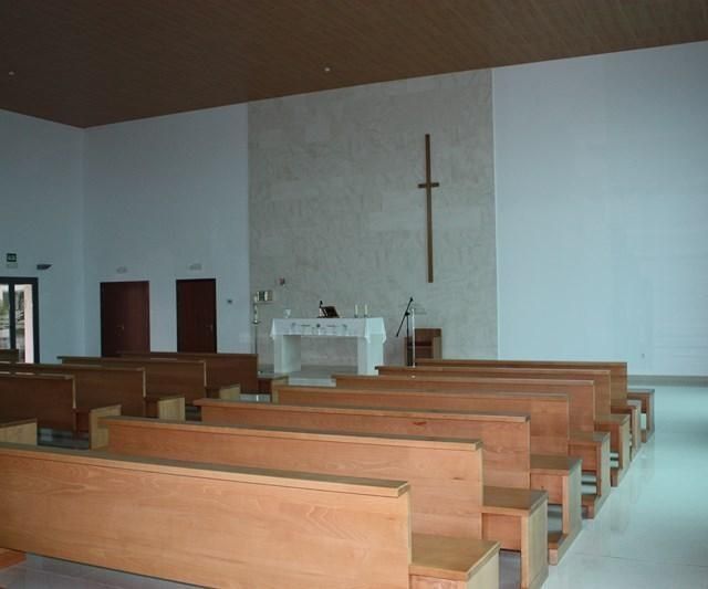 Interior vacío de la iglesia con bancos de madera, altar y una cruz en la pared frontal.