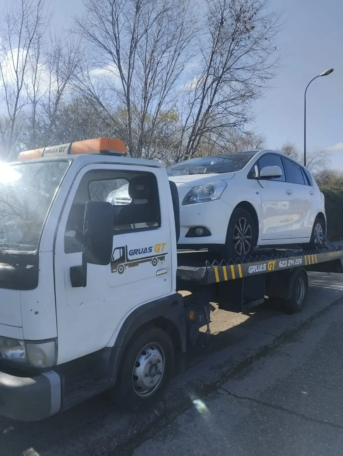 Automóvil blanco sobre una grúa, estacionado en una calle con árboles y un poste de luz.