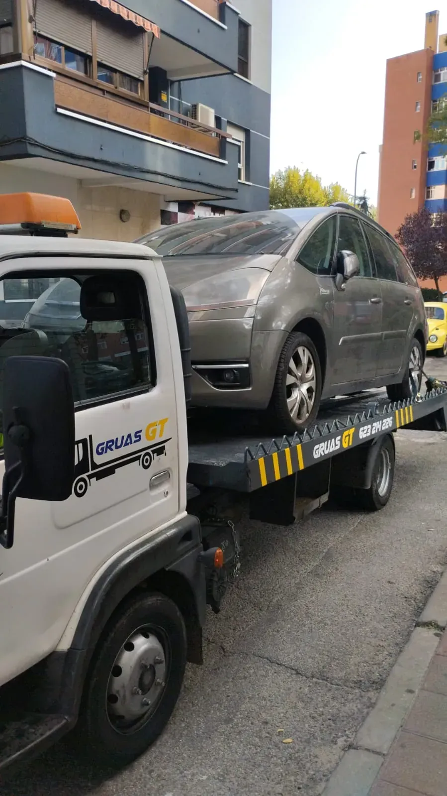 Grúa que transporta un Citroën Picasso gris en una calle de la ciudad.
