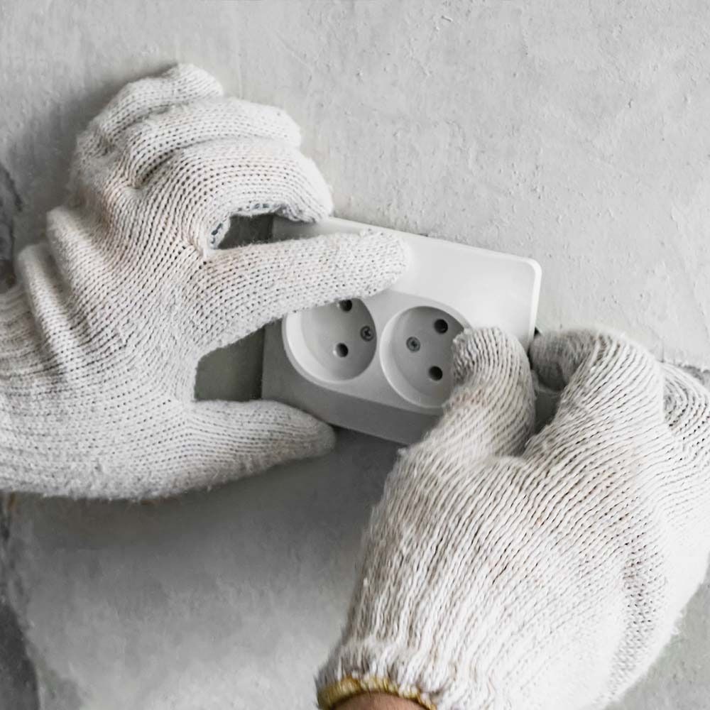 Manos con guantes blancos instalando una toma de corriente blanca en una pared gris.