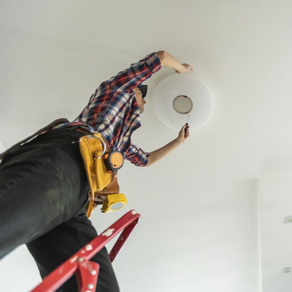 Electricista en una escalera instalando una lámpara de techo en una habitación blanca.