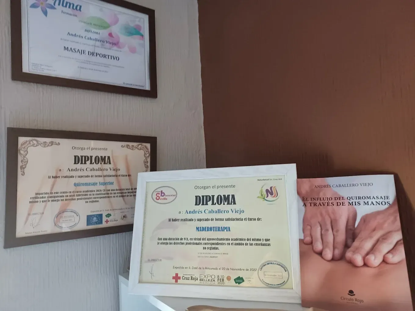 Certificados y libro en la pared: tres diplomas enmarcados y un libro con una ilustración de masaje.