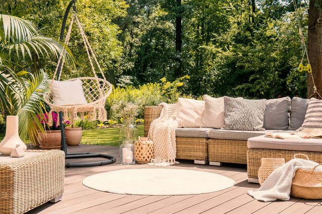 Patio extérieur confortable avec mobilier en osier, coussins crème, table basse ronde et jardin verdoyant en arrière-plan.
