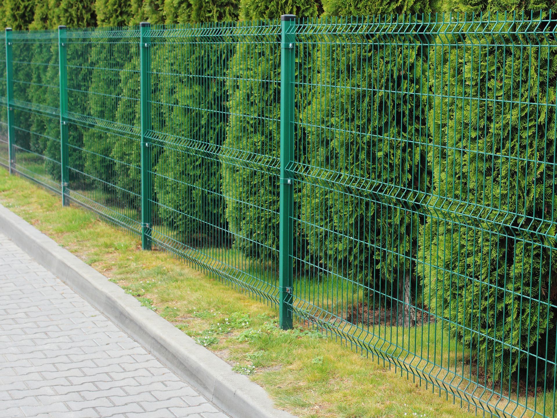 Une clôture métallique verte longe un trottoir pavé, bordé de haies denses à l'arrière.