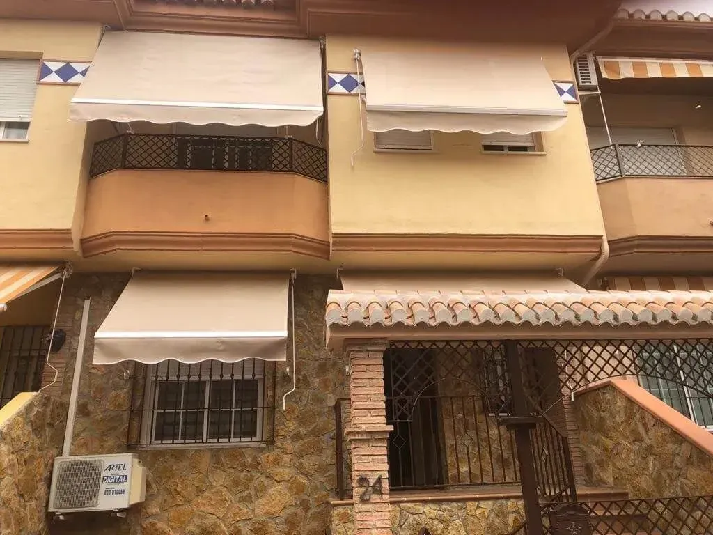 Vista frontal de un edificio de apartamentos de color beige con toldos, ventanas con rejas y una entrada con verja.