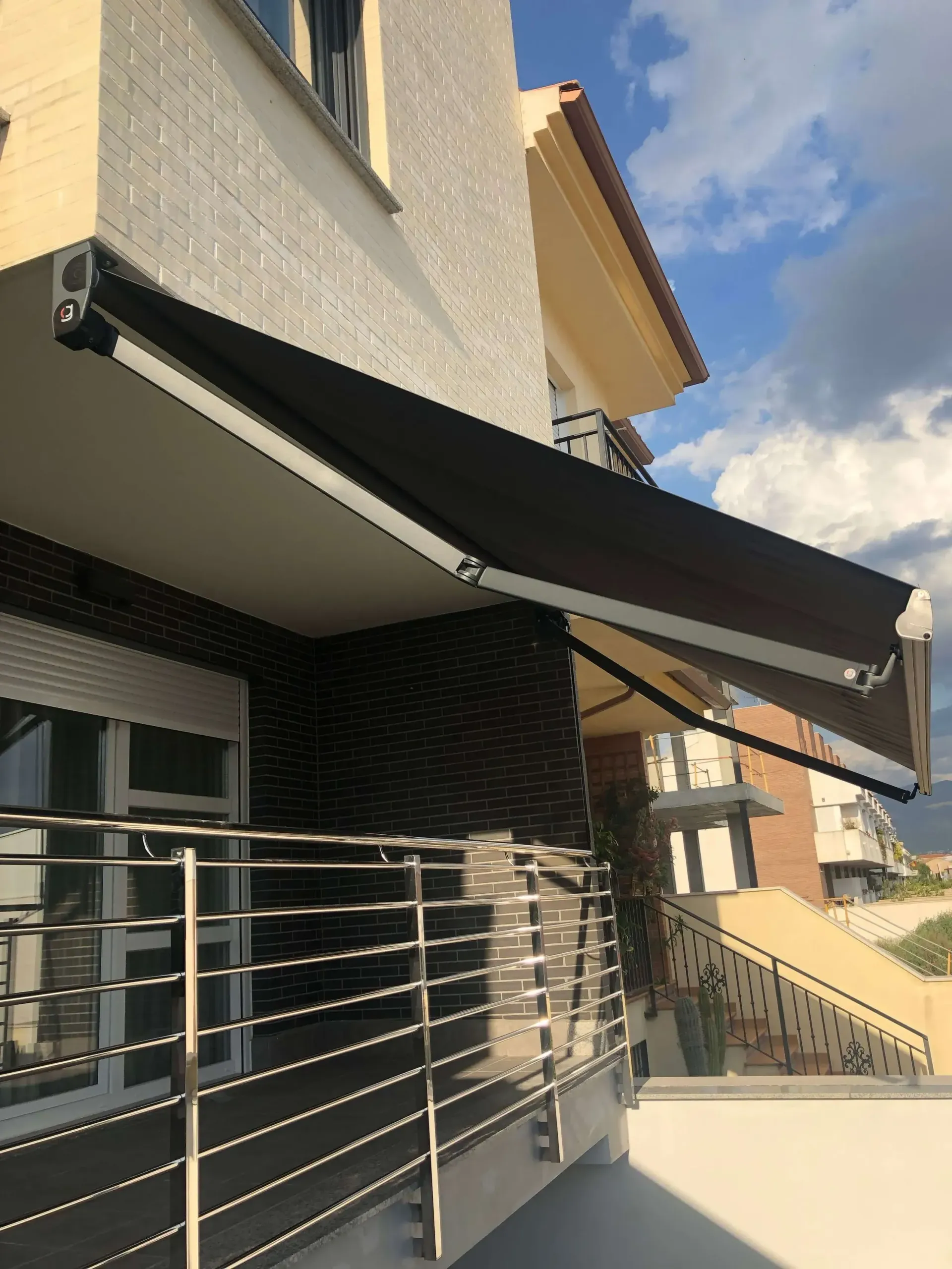 Toldo retráctil negro sobre la entrada de un edificio con barandilla metálica bajo un cielo azul.