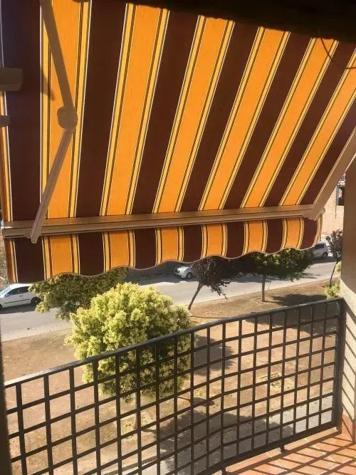 Toldo a rayas naranjas y blancas sobre un balcón, con vistas a una calle y a un árbol frondoso.