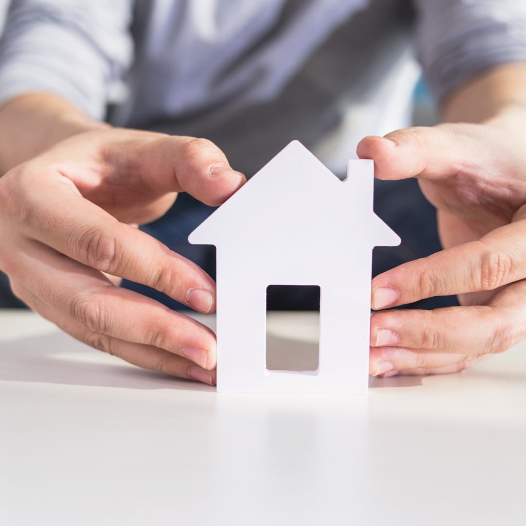 Hands holding a white paper cutout of a house on a white surface.