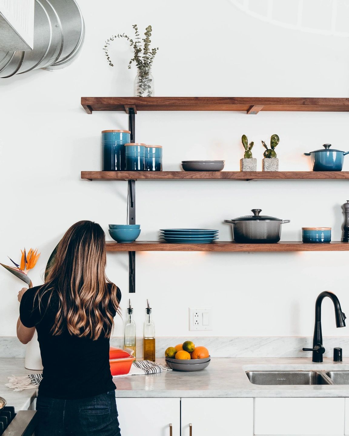 A person stands at a kitchen counter with open wooden shelving holding blue kitchenware, potted cacti, and decor.