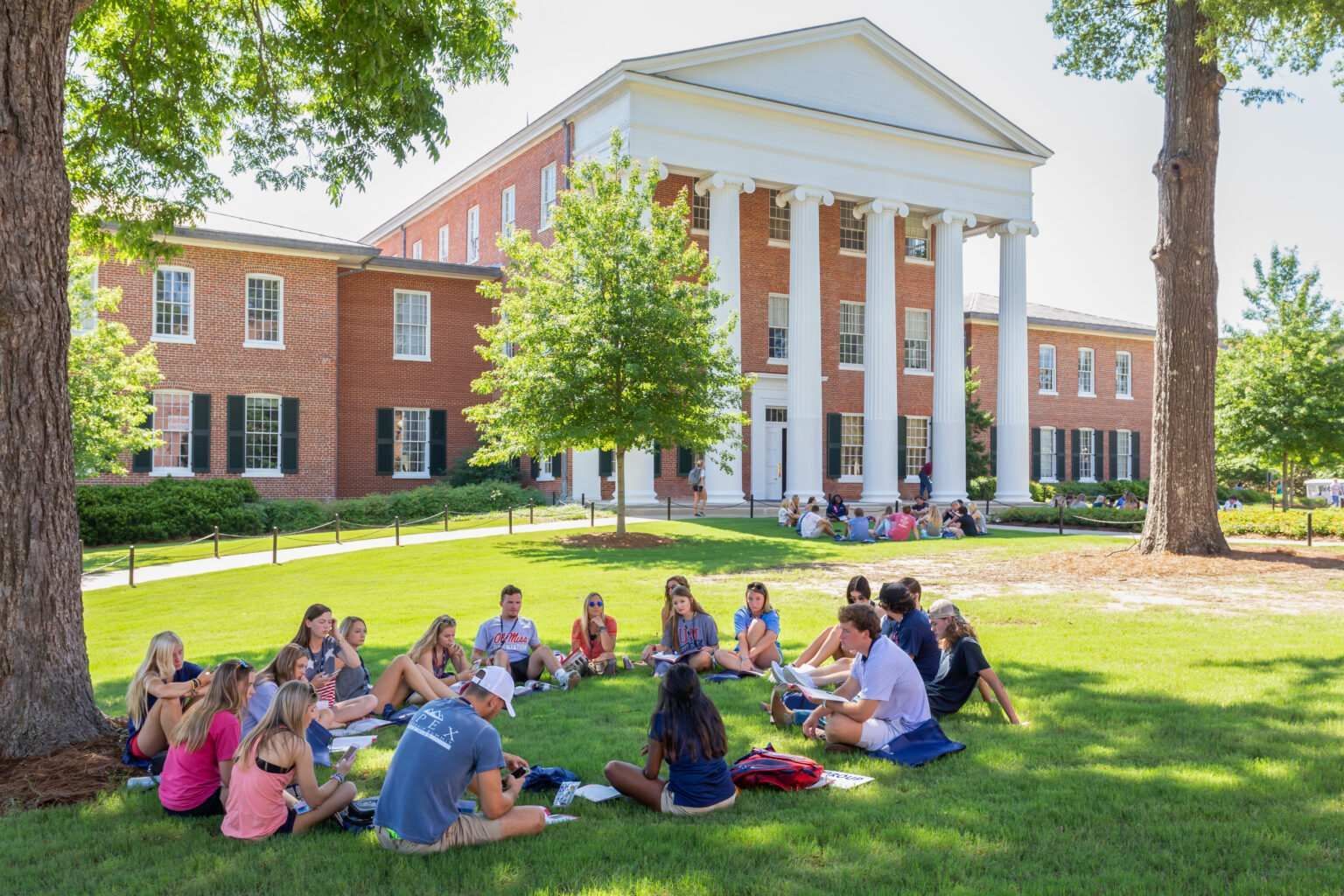 A group of students sit in a circle on a grassy lawn in front of a large, red-brick building with white columns.