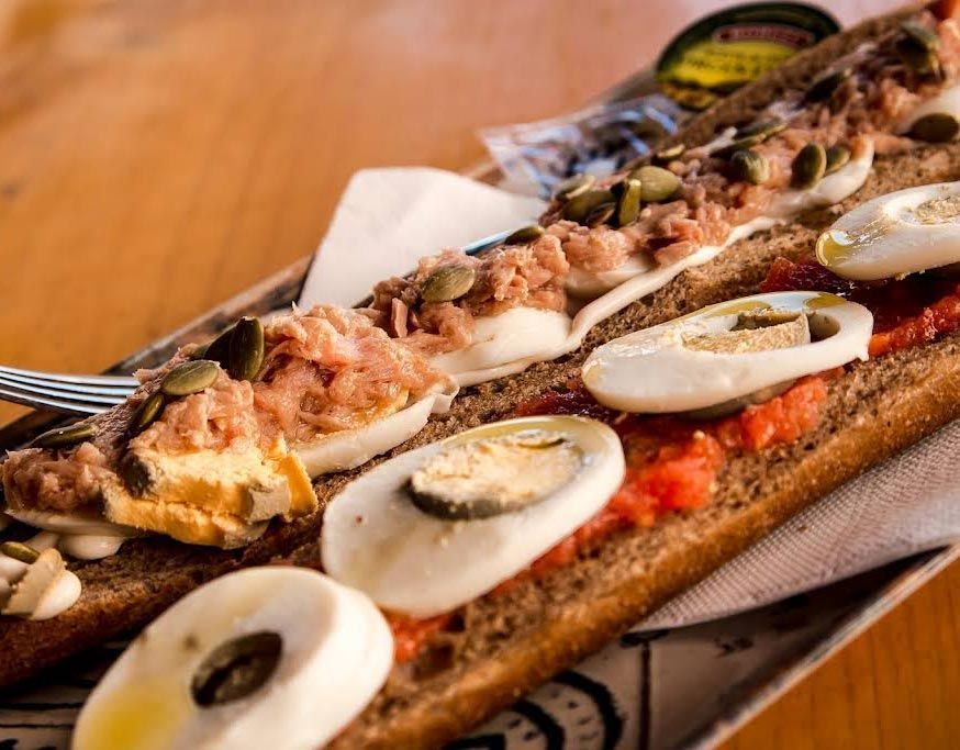 Un sándwich largo con atún, huevos y tomates sobre una mesa de madera.