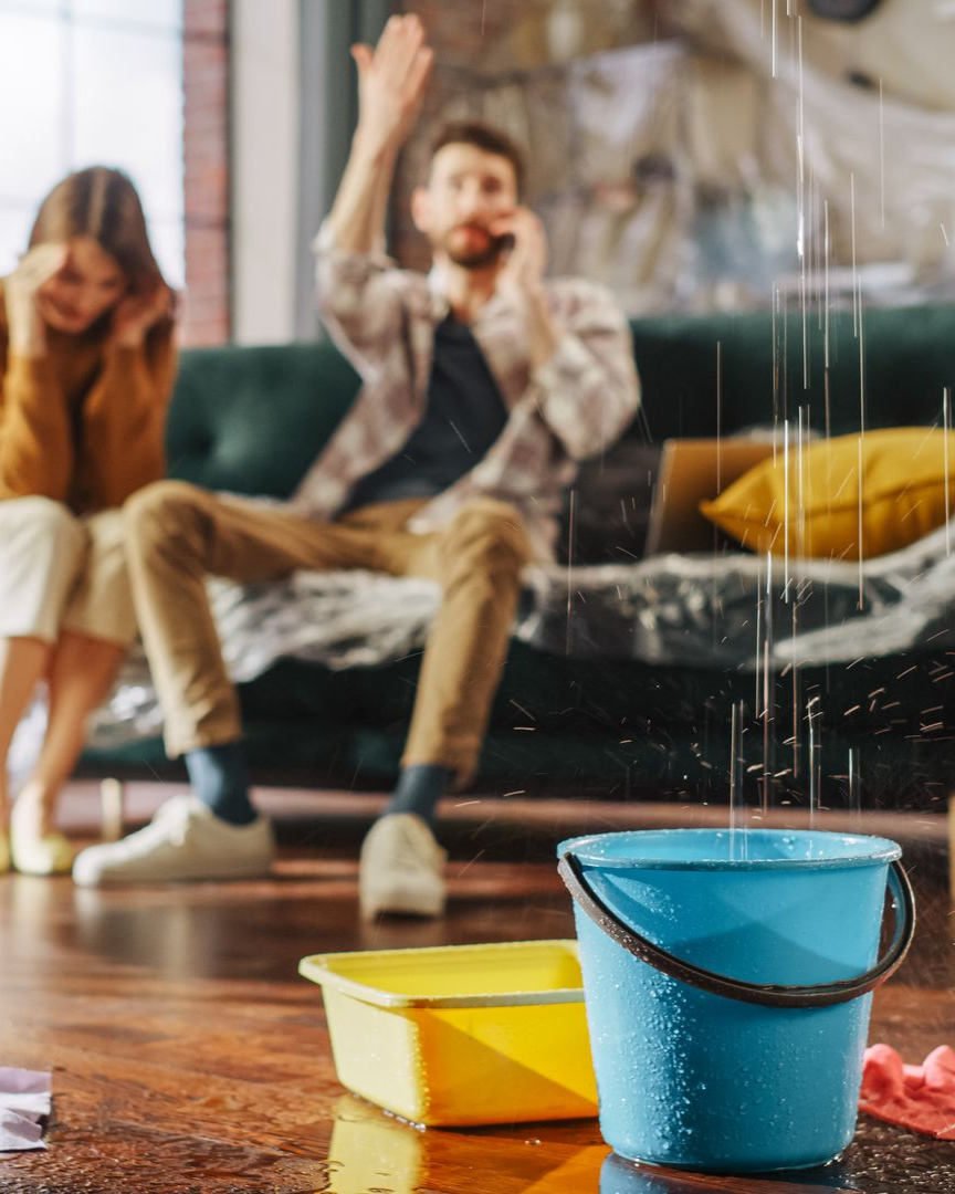De l'eau s'infiltre du plafond dans un seau, avec des personnes au téléphone en arrière-plan.
