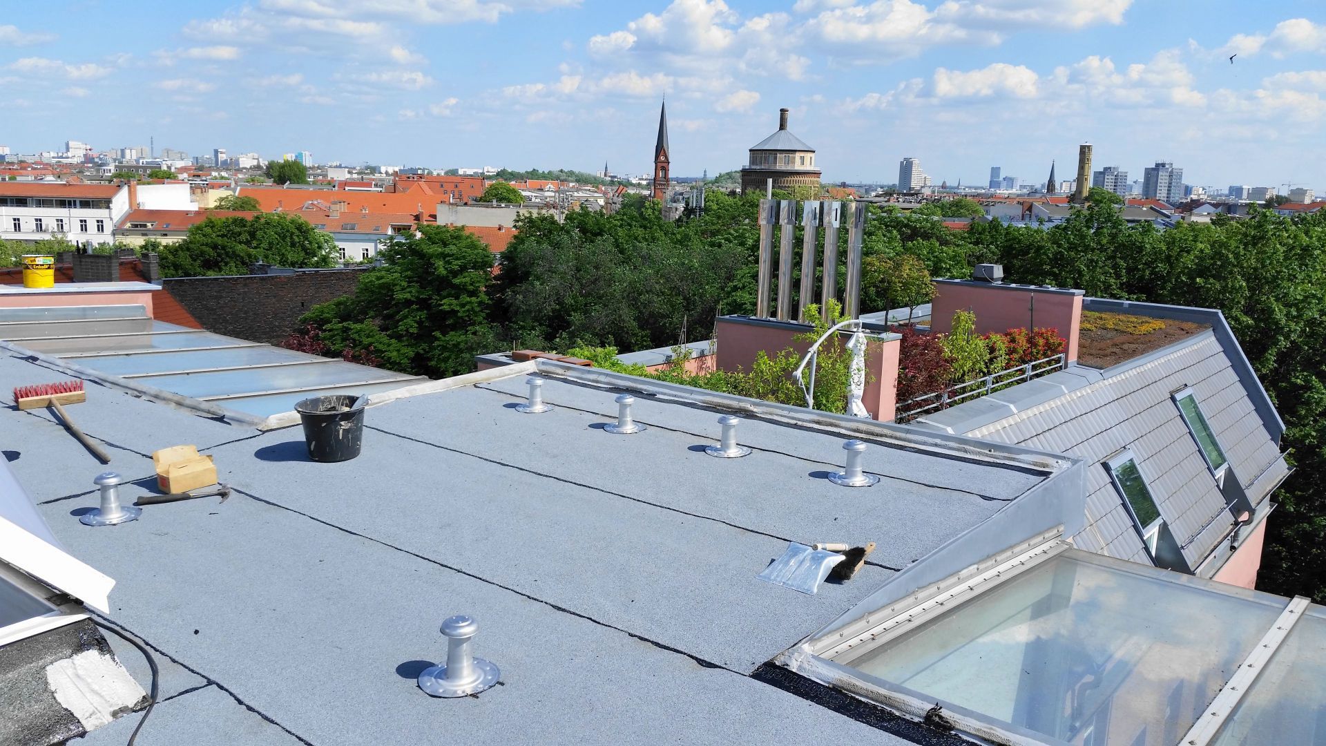 Ein Flachdach in Reparatur, mit grauer Membran, Lüftungsrohren und Oberlichtern, im Hintergrund eine Stadtkulisse.