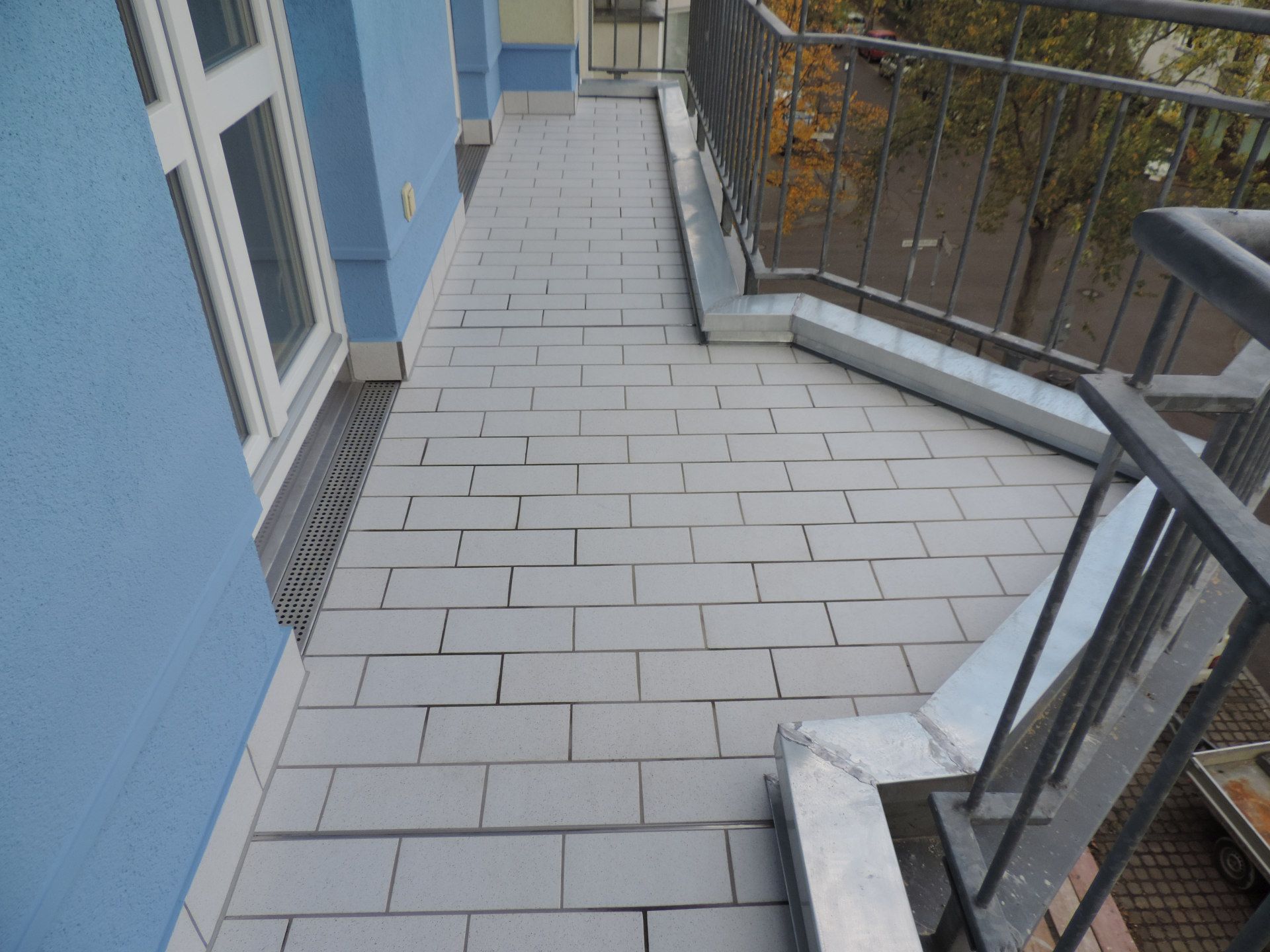 Ein schmaler Balkon mit hellgrauen Fliesen neben einer blauen Gebäudewand und einem Metallgeländer.
