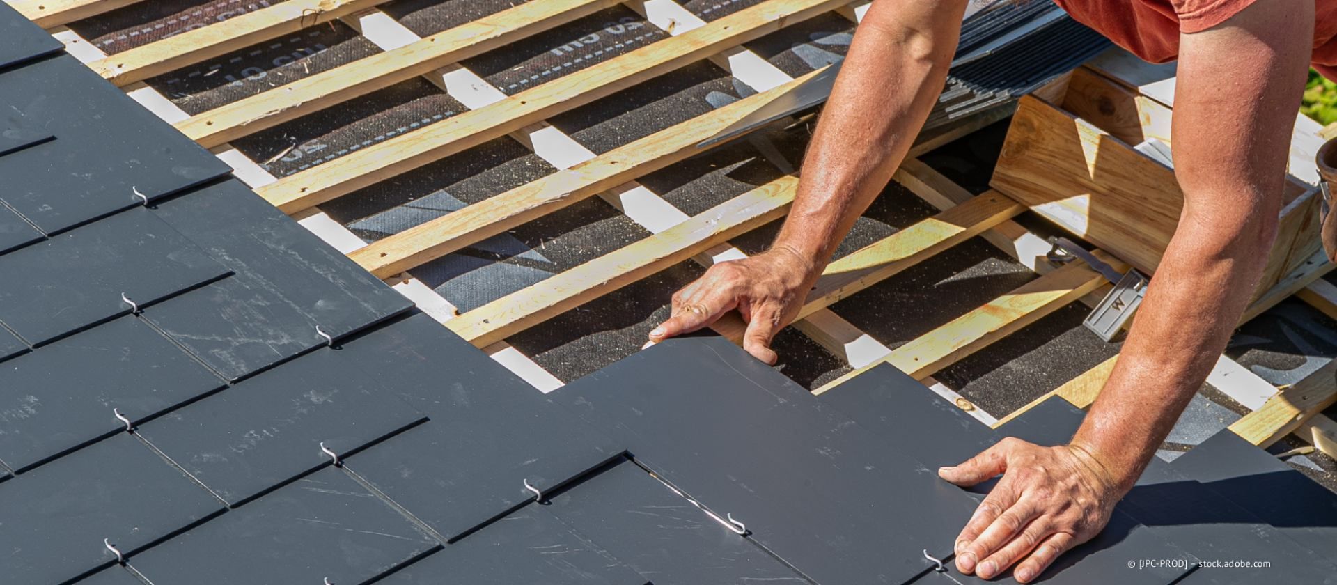 Eine Person verlegt dunkelgraue Dachziegel auf einem Holzgerüst.