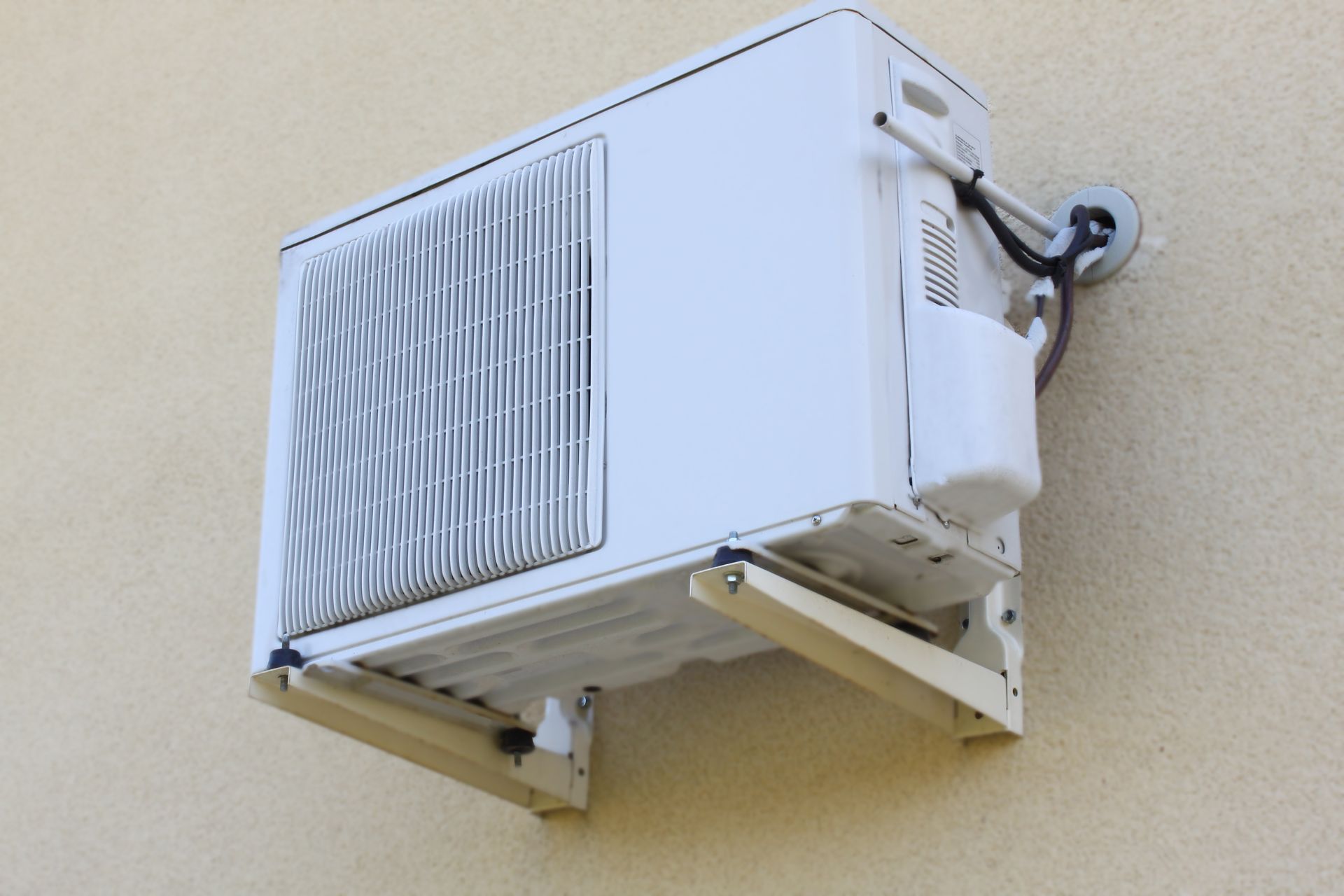 Climatisation extérieure blanche montée sur un mur beige avec supports de fixation.