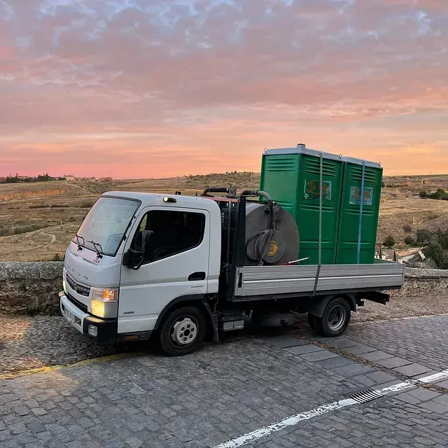 Un camión blanco con baños portátiles verdes en la parte trasera está estacionado en una calle adoquinada.