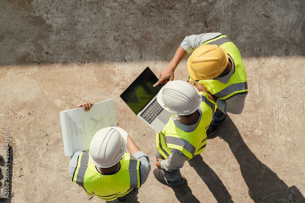 Bauarbeiter in Sicherheitswesten und Schutzhelmen studieren Baupläne und Laptops auf einer Betonfläche.