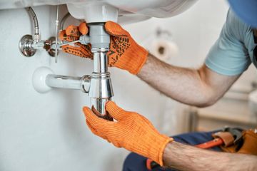 Un fontanero con guantes de trabajo naranjas instala un sifón cromado debajo del lavabo de un baño.