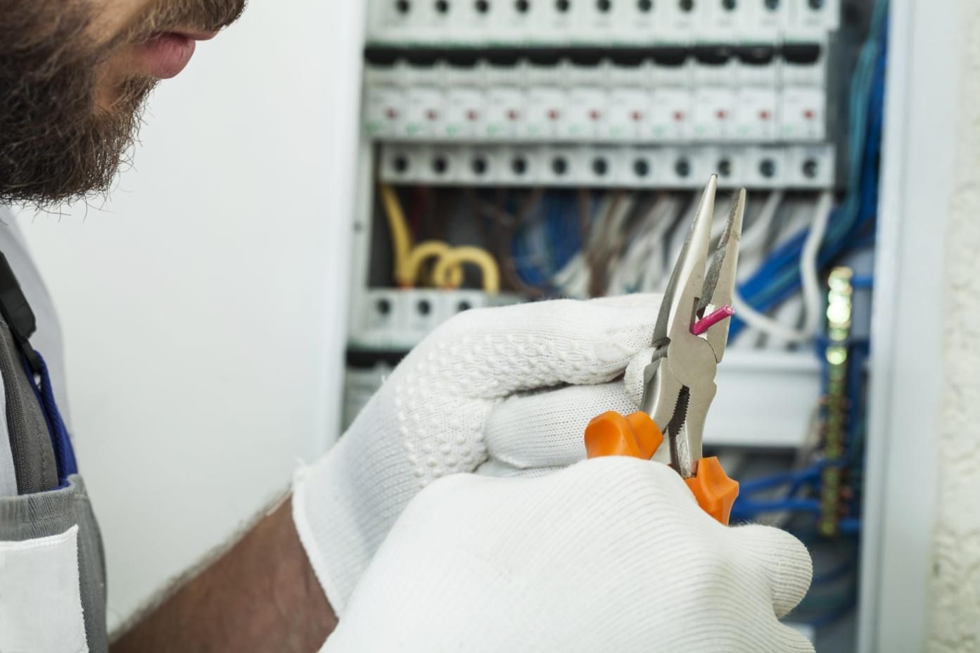 Un electricista, con guantes blancos, utiliza unos alicates para manipular los cables dentro de un panel eléctrico.