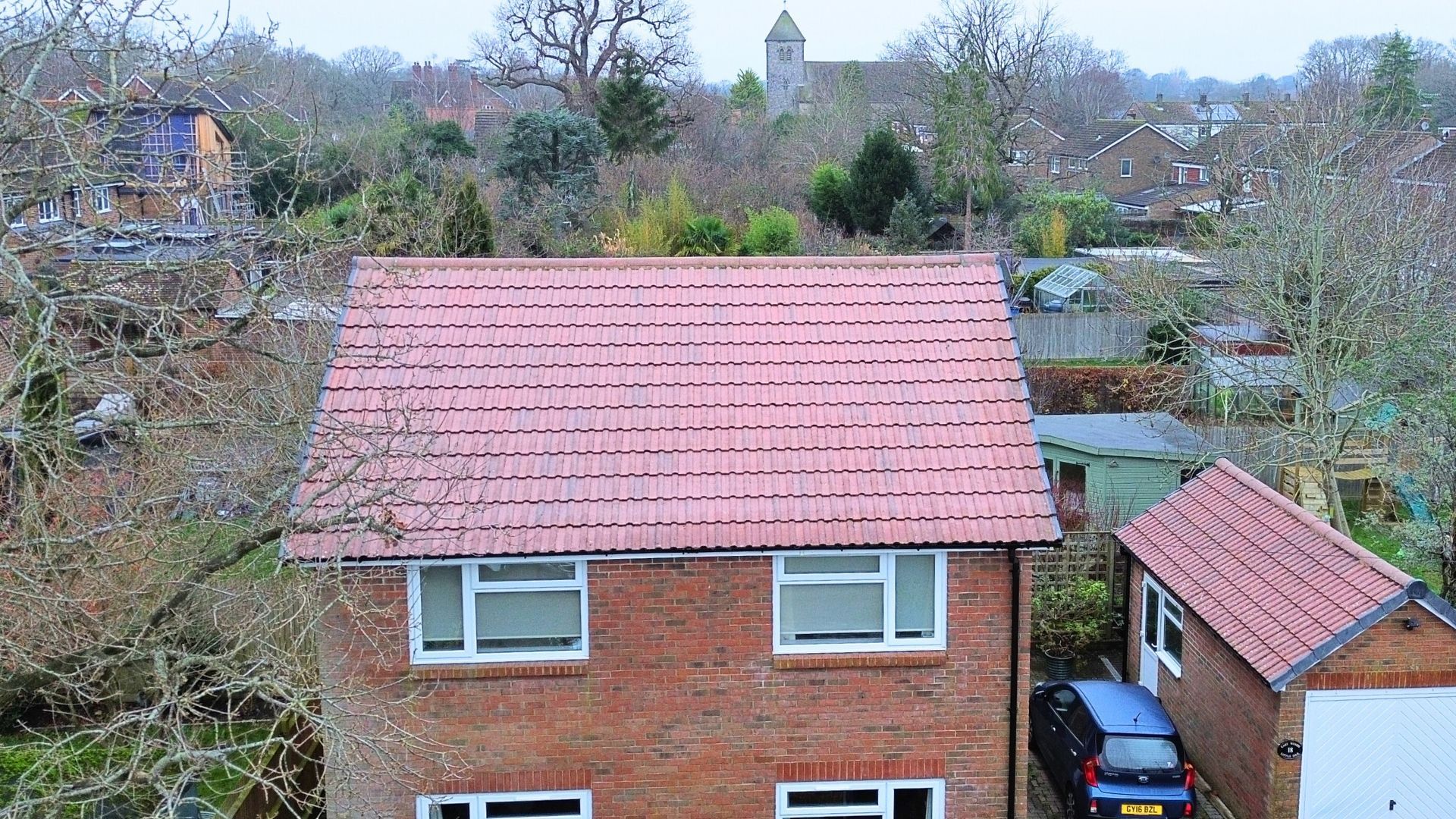 Brick house with red roof and white windows beside a small garage, viewed over a neighborhood.