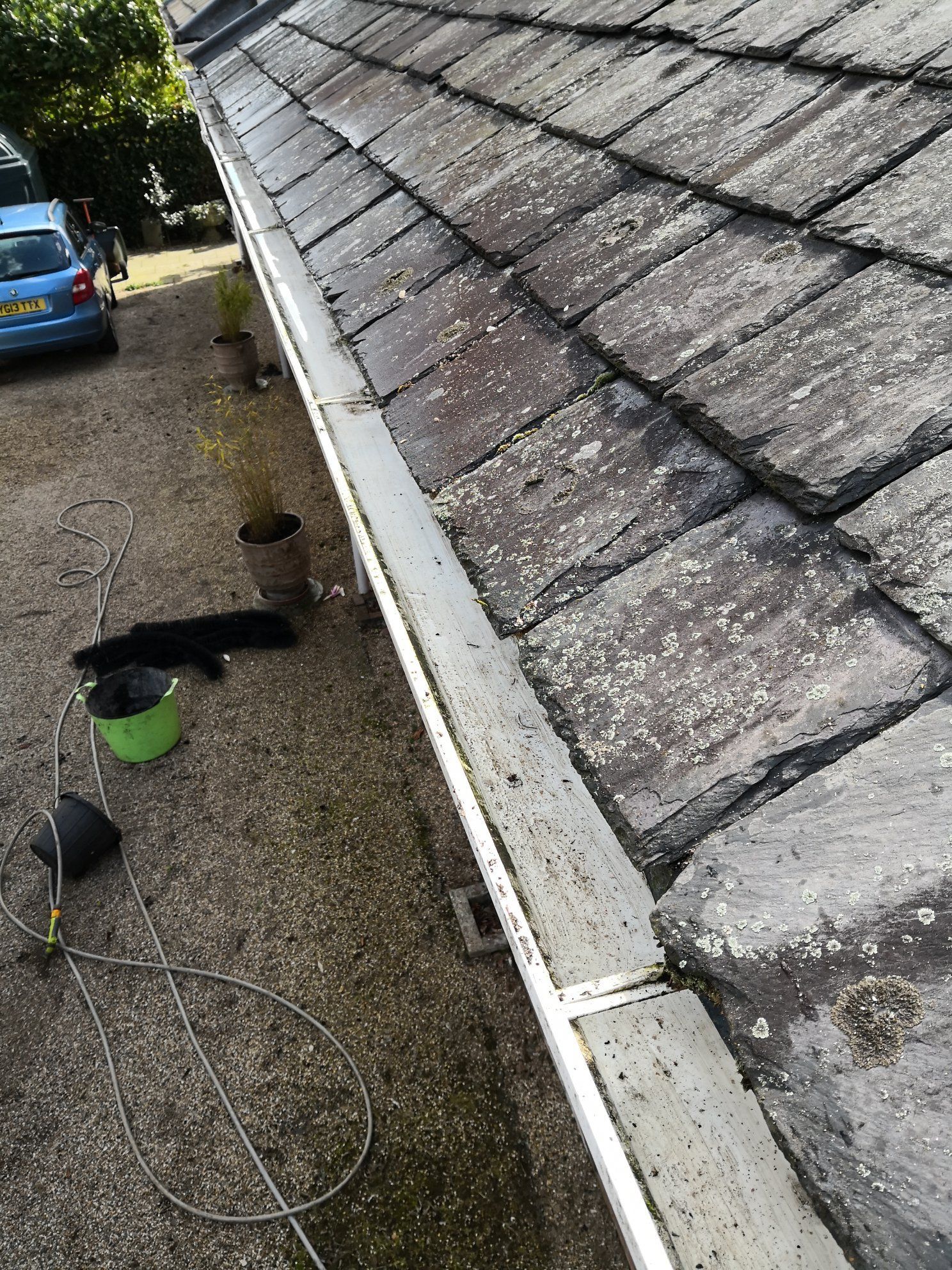 Roof shingles and gutter along a house eave, with gravel below and a blue car in the background.