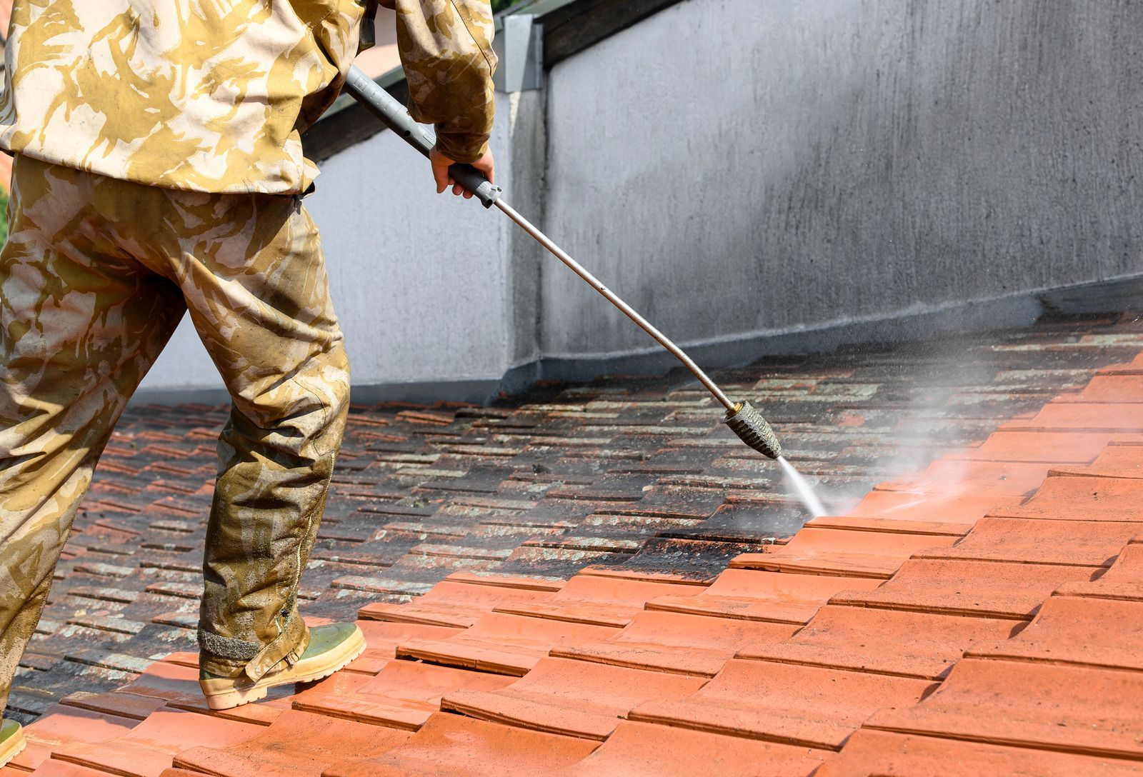 Worker pressure-washing a red brick roof beside a gray wall