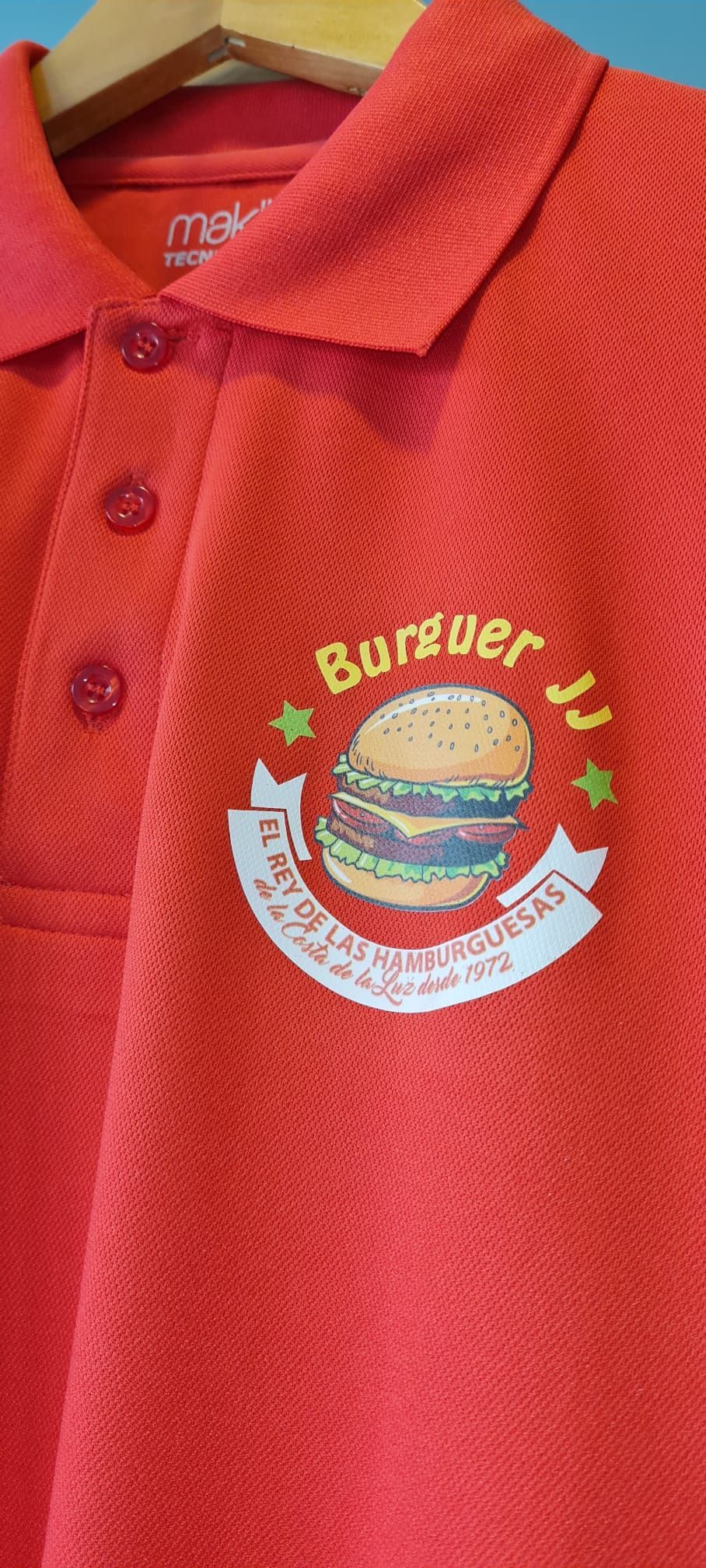 Un polo rojo con una hamburguesa bordada cuelga de una percha.