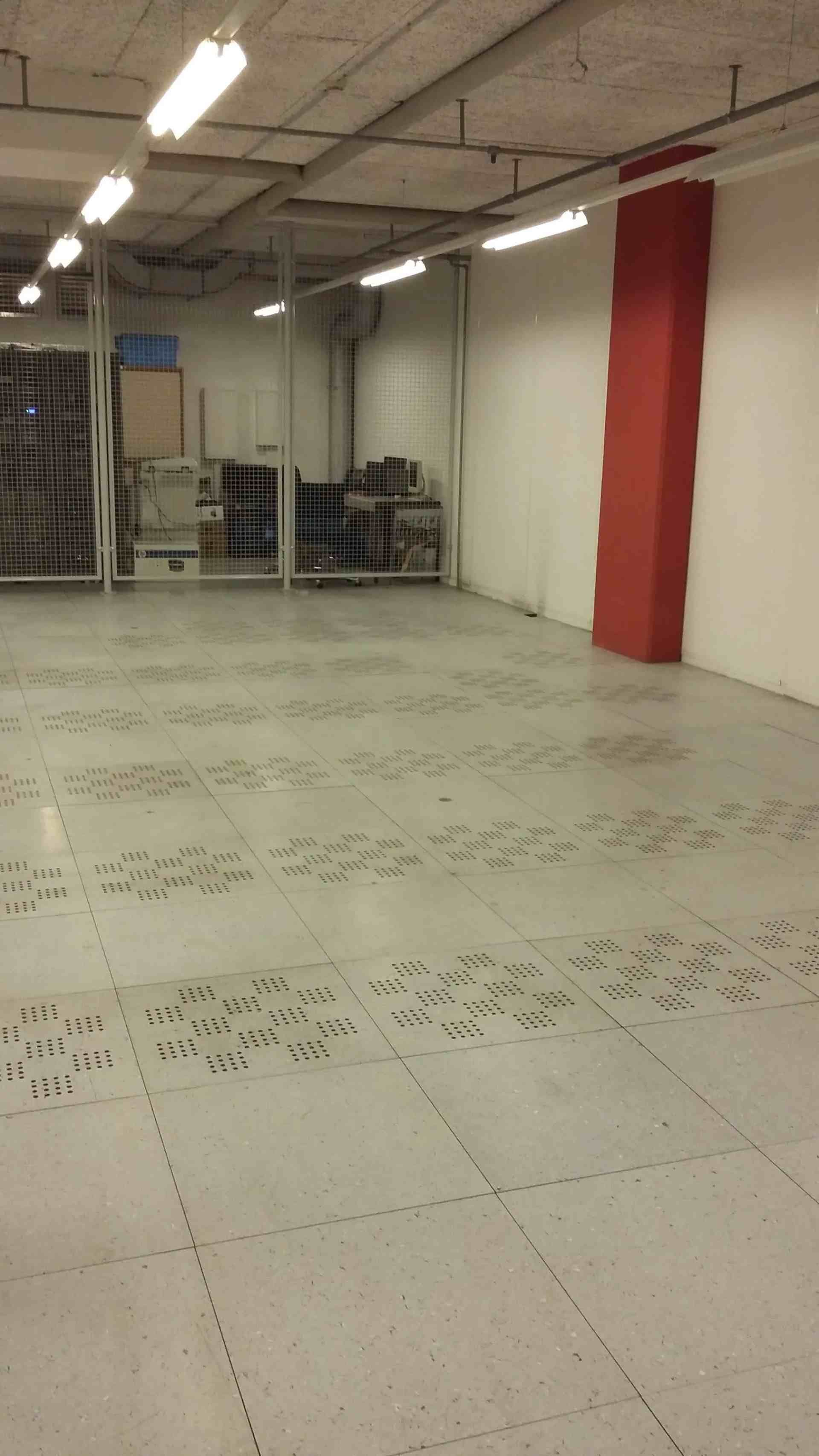 Intérieur d'entrepôt vide, sol carrelé gris, pilier rouge, étagères en arrière-plan. Éclairage fluorescent au plafond.