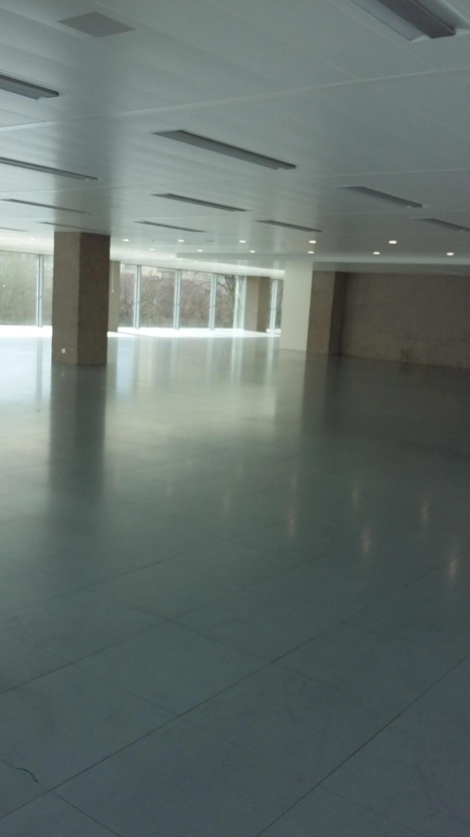 Grande pièce vide avec sol réfléchissant et longs luminaires au plafond. Fenêtres le long du mur du fond.