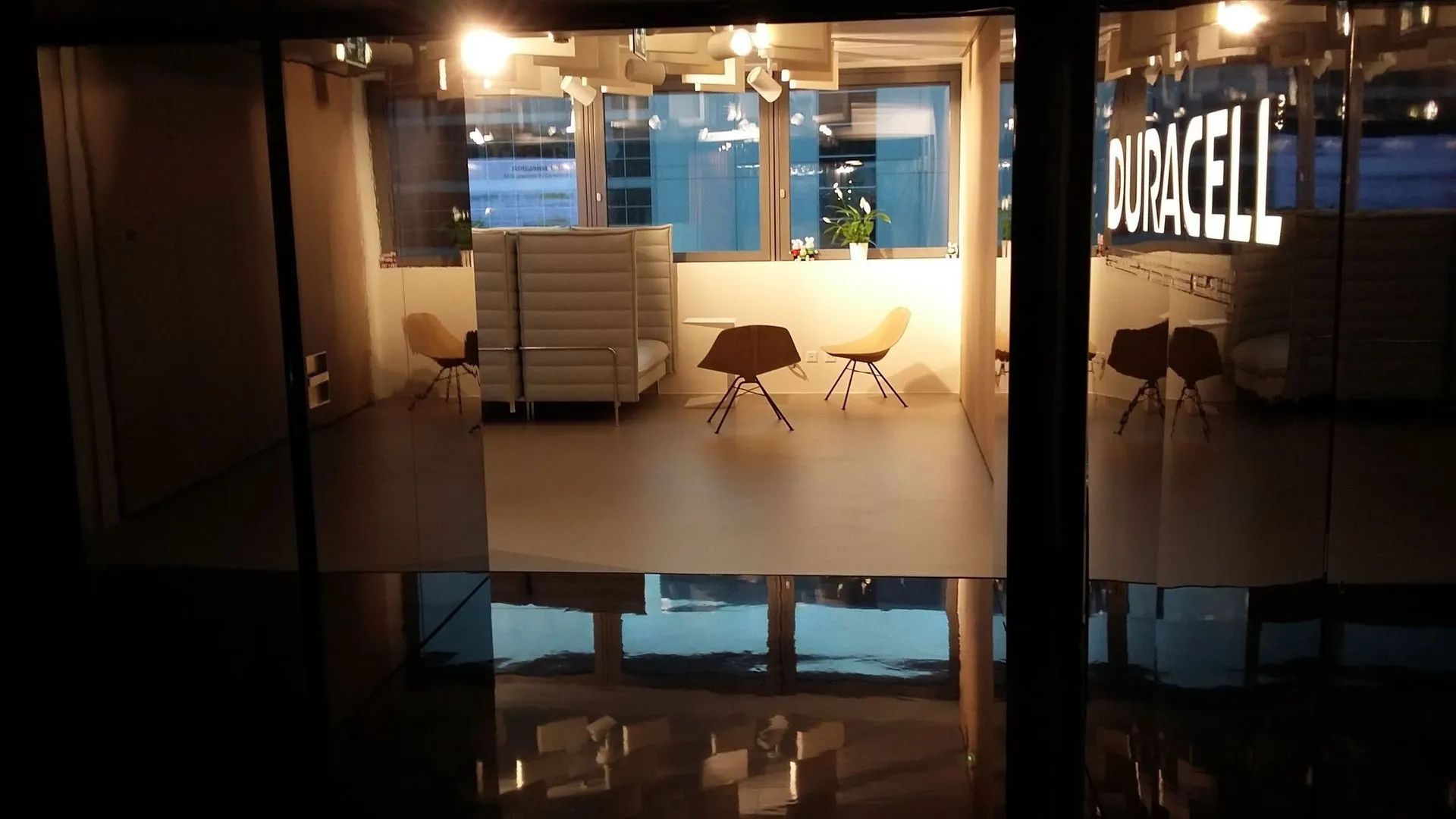 Vue à travers une vitre sur un espace de bureaux. La pièce est lumineuse, avec des sièges et une enseigne Duracell.