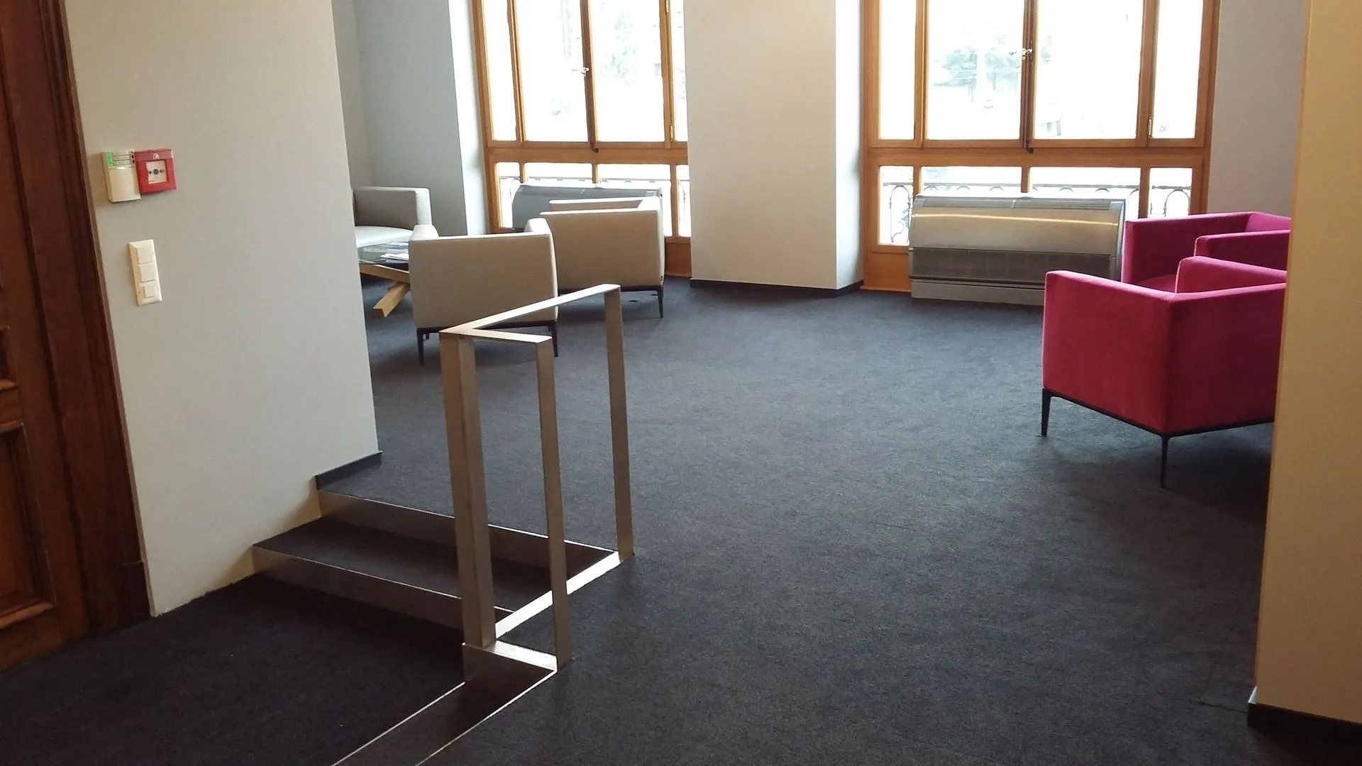 Intérieur avec escalier, moquette grise, coin salon avec fauteuils roses et gris, fenêtres.