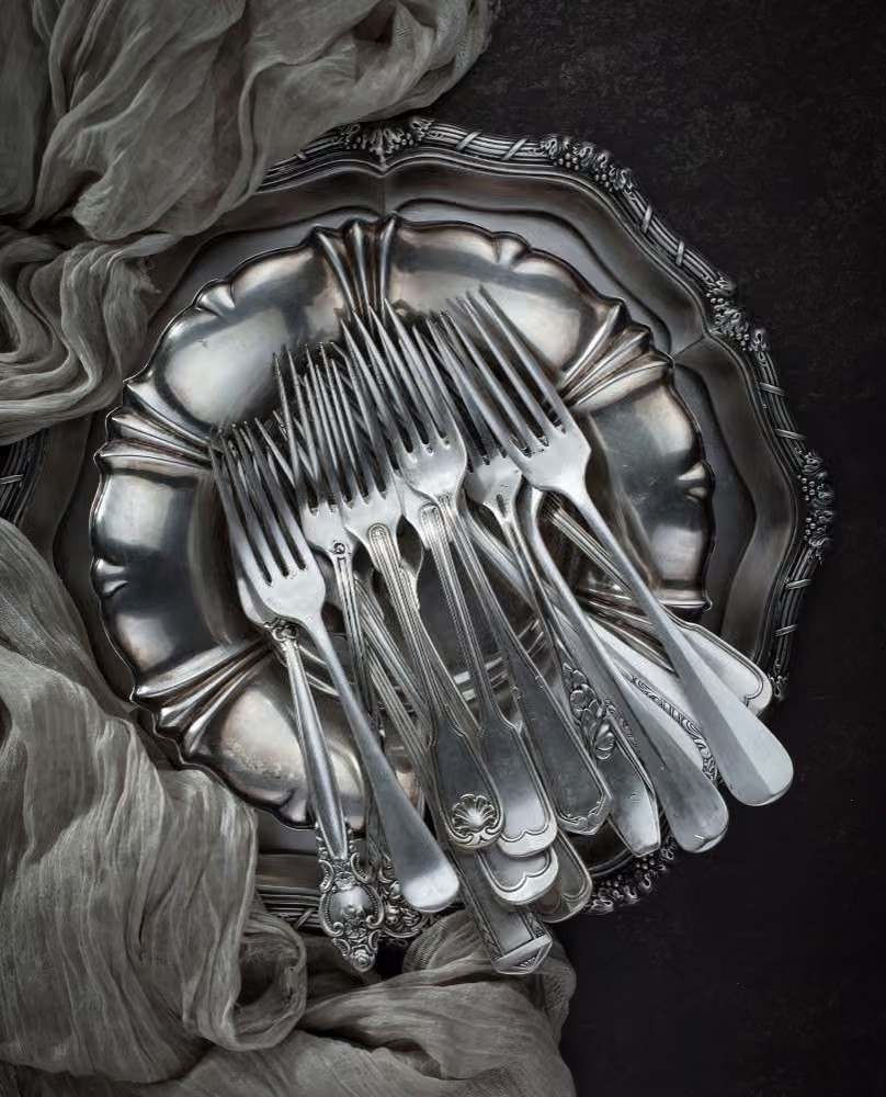 Tenedores y cucharas de plata cuidadosamente dispuestos en una bandeja ornamentada con un drapeado de tela gris.