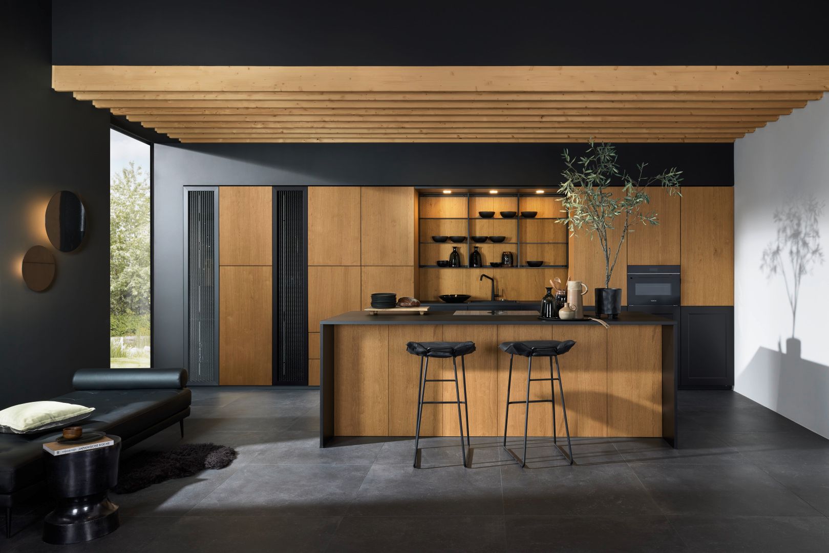 Présentation de la cuisine aux tons bois et noir dans un espace éclairé assorti avec îlot central bi-ton noir et bois et 2 tabourets noirs.