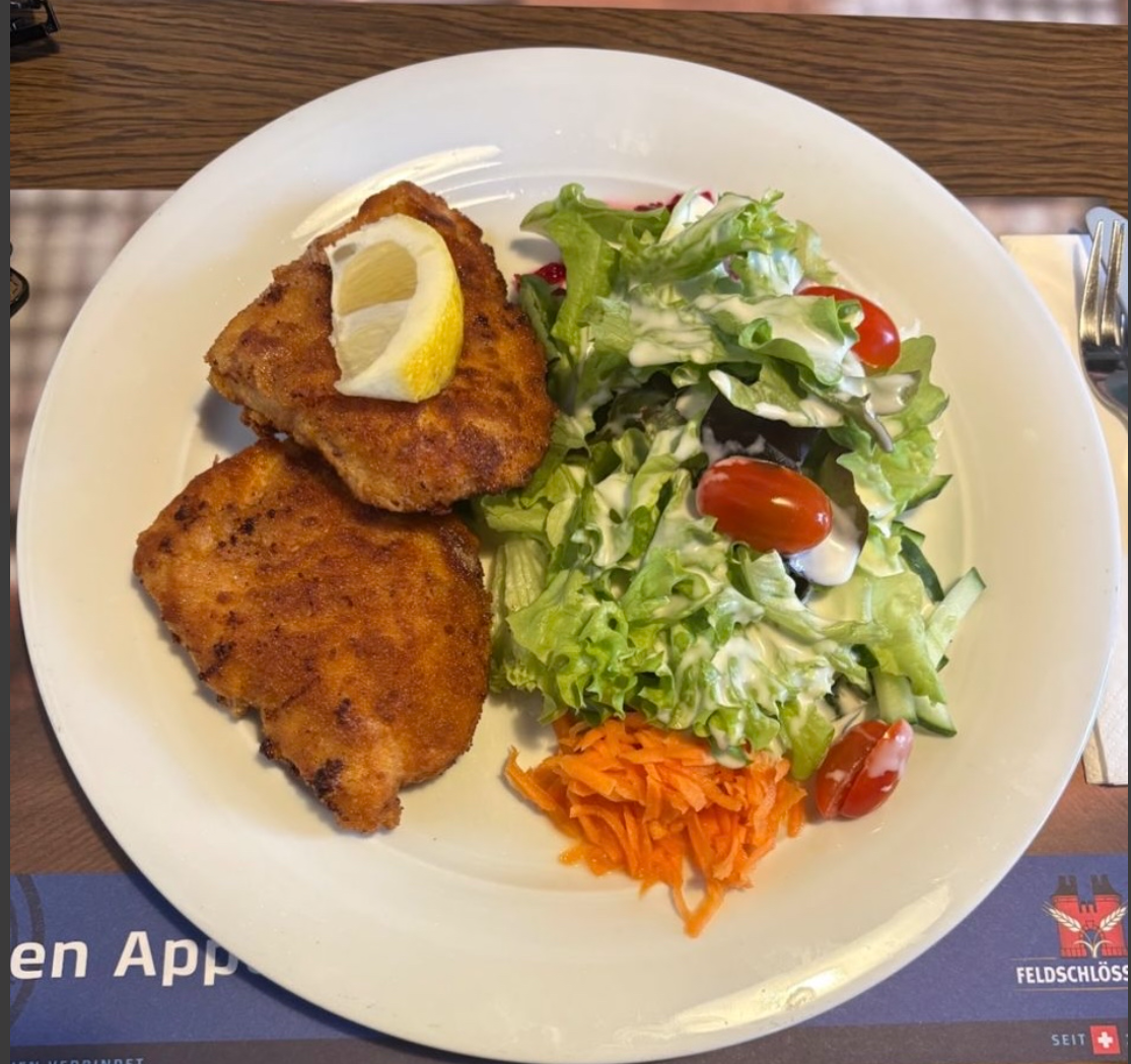Panierte Schnitzel auf einer schwarzen Pfanne mit Zitrone, Kräutern und Sauce, dazu zwei Marmeladengläser auf einem Holzbrett.