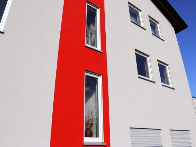 Extérieur d'un bâtiment moderne avec des murs gris, des accents rouges et des fenêtres à cadre blanc sous un ciel bleu.