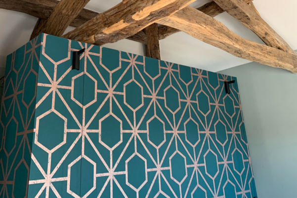 Mur à motifs géométriques bleu sarcelle avec des lignes roses, contre un plafond blanc avec des poutres en bois apparentes.