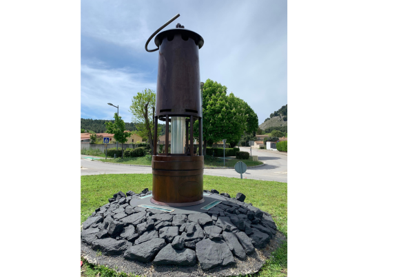 Monument d'une lampe de mineur cylindrique sur une base de charbon; situé à l'extérieur avec des arbres et le ciel.