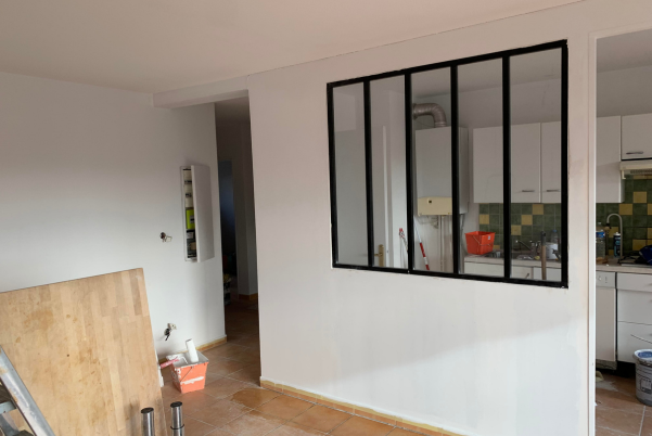 Vue intérieure : un mur avec une fenêtre à cadre noir donnant sur une cuisine ; un couloir est visible à gauche.