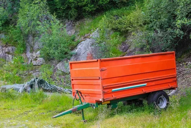 Remolque agrícola naranja en un campo de hierba con un fondo verde de árboles y rocas.
