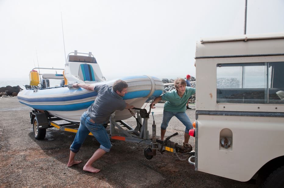 Dos personas empujan una embarcación sobre un remolque, conectada a una camioneta blanca, en una zona costera.