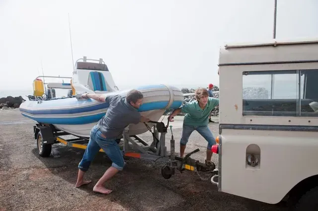 Dos personas empujan un bote en un remolque, conectado a un vehículo blanco, en una rampa para botes.