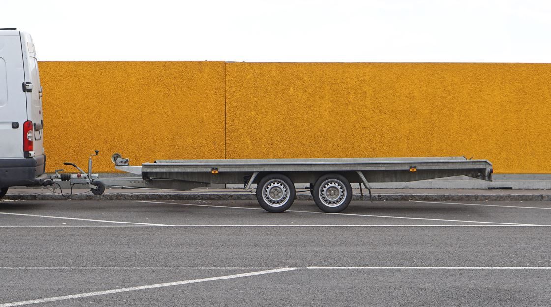 Furgoneta blanca remolcando un remolque de plataforma sobre asfalto junto a una pared naranja.