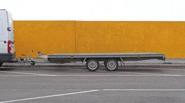 Furgoneta blanca remolcando un remolque de plataforma largo y vacío en una calle con una pared amarilla al fondo.