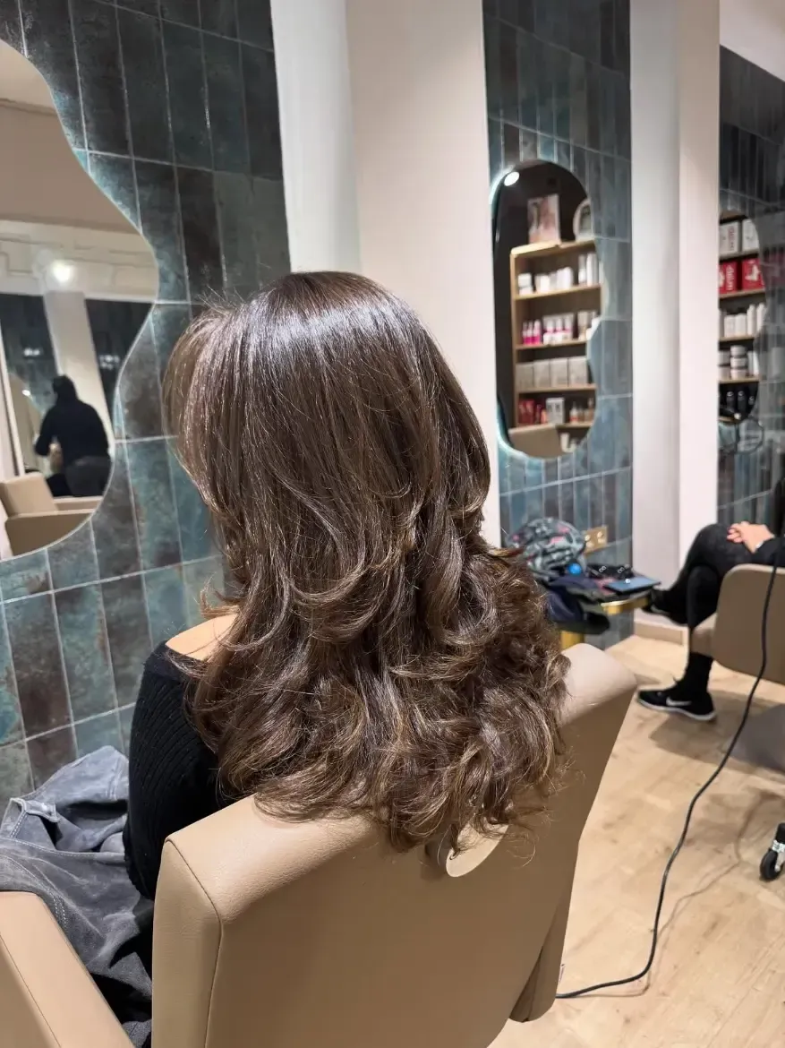 Clienta de peluquería con cabello castaño largo y ondulado, sentada en una silla beige con un espejo