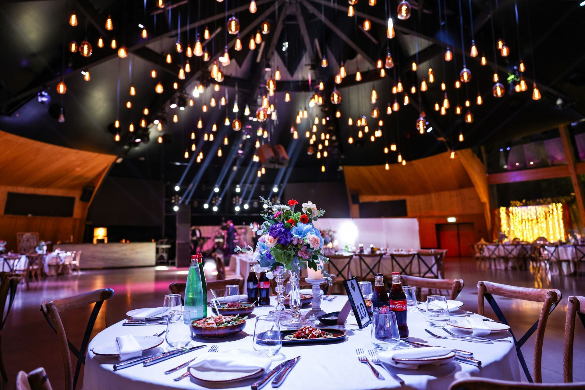 Elegante salón de banquetes con mesa redonda, centro de mesa floral y guirnaldas de luces colgantes.