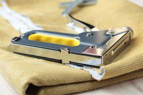 Grapadora sobre tela color canela, grapando material blanco. Herramienta plateada y amarilla.