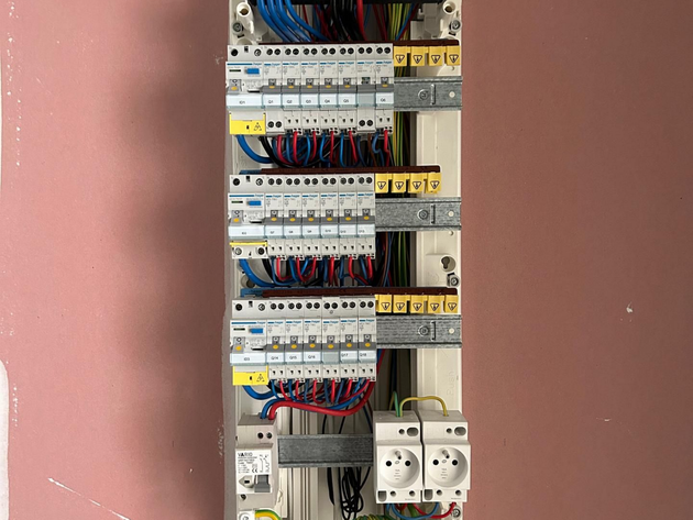 Tableau électrique avec des rangées de disjoncteurs et de câbles sur un mur rose.