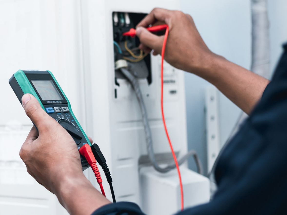 Une personne utilise un multimètre pour tester des fils électriques à l'intérieur d'un panneau blanc.
